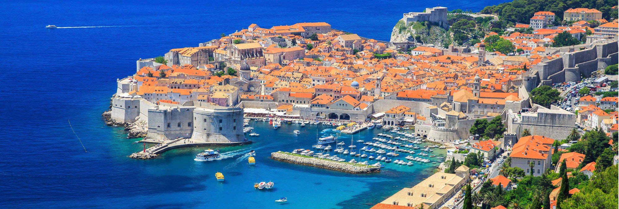 Historic coastal town with red-tiled roofs, medieval stone walls, and turquoise harbor filled with boats on Adriatic Sea